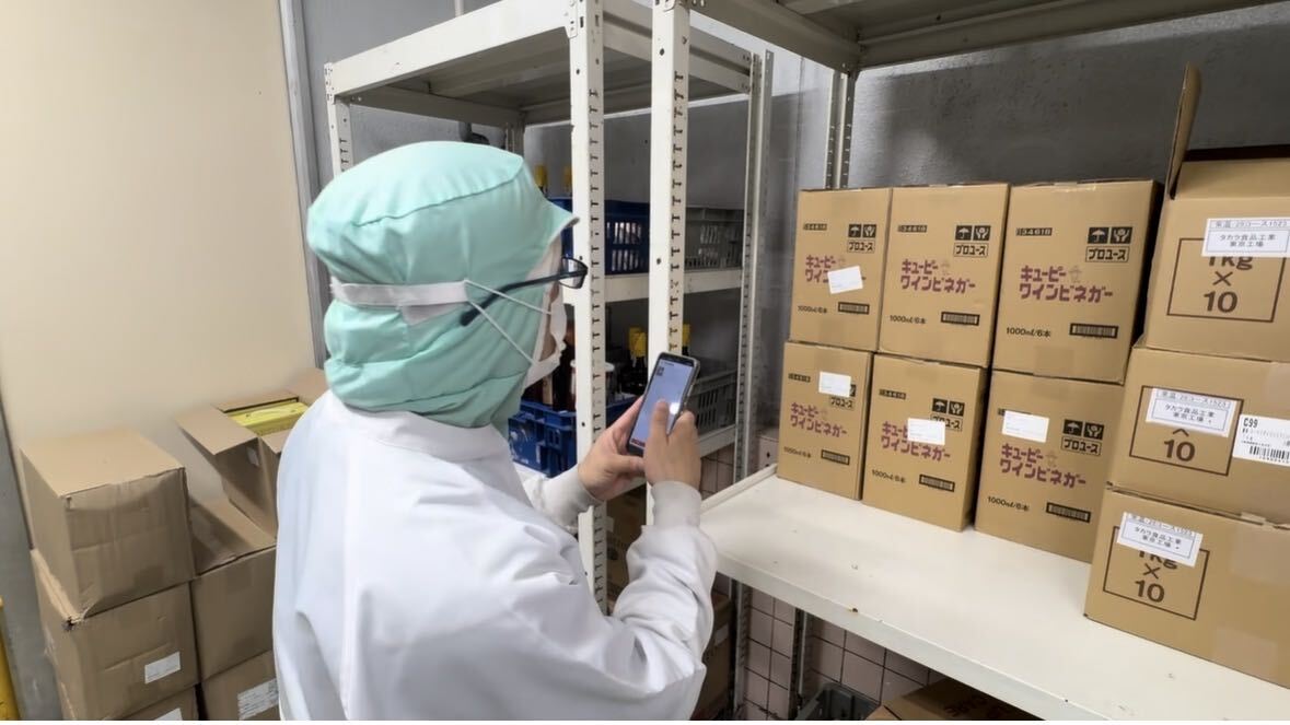 食品工場の棚の前で、作業服のままスマートフォンで商品を撮影している様子